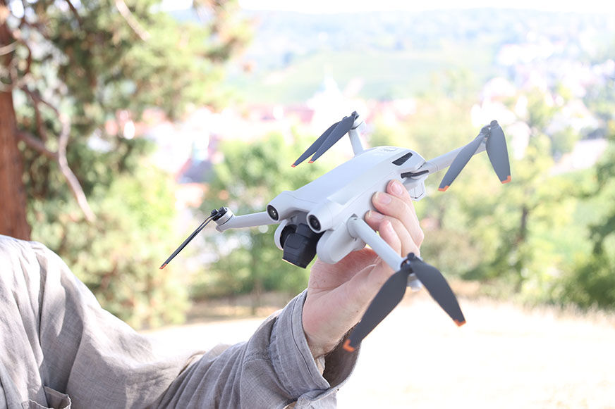 Die DJI Mini 3 Pro wird in der Hand gehalten und ist hellgrau und die Rotorblätter sind schwarz mit einem Orangestich.